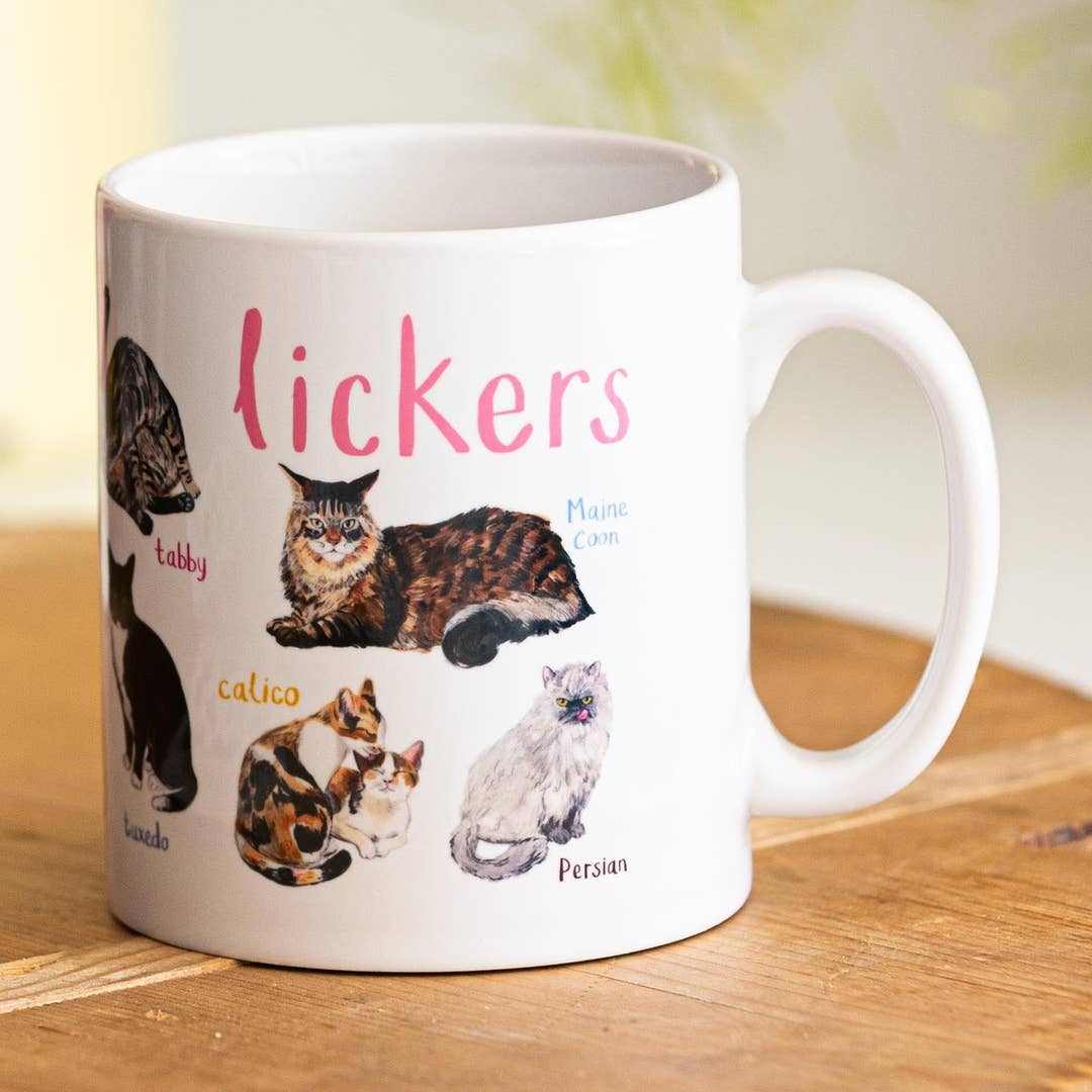 A mug with cat illustrations on a white background. The text 'Lickers' is printed over the illustrations along with text labelling each image. The mug sits on a wooden surface.