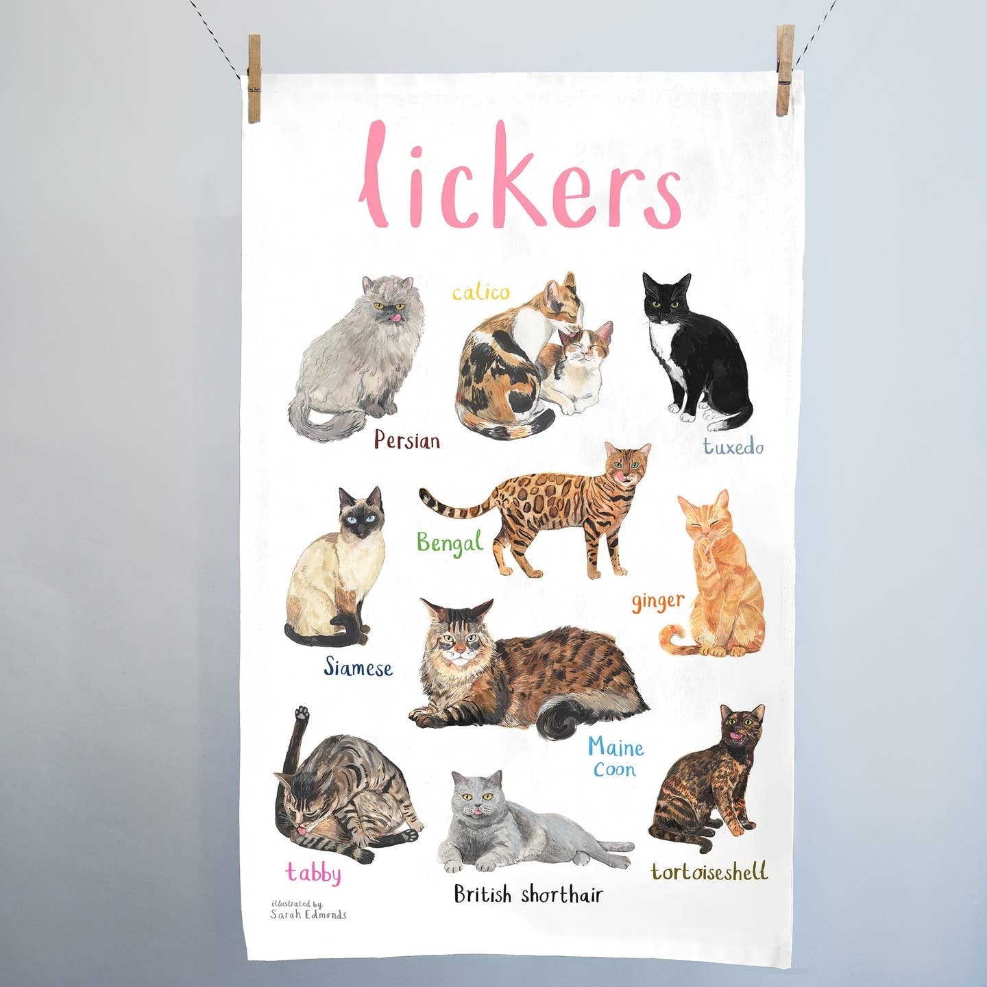 A white cotton tea towel showing colourful cat illustrations with text  printed underneath. The heading 'lickers' is printed at the top. The tea towel hangs from 2 clips on a string. 