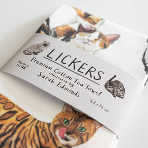 A white cotton tea towel showing cat illustrations with text printed underneath. The tea towel is folded and a paper label titled 'Lickers' is wrapped around the middle.