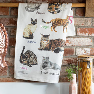 A white cotton tea towel showing cat illustrations with text printed underneath. The tea towel is folded and hangs from a wooden dowel against a brick wall background. 