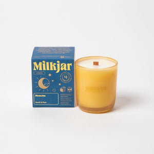 Canadian company Milkjar candle and its box packaging. The white candle is in an yellow glass with "Milkjar" written in gold. The other side of the candle has a cute smiling moon-sun-hybrid doodle in gold. The packaging is a small navy blue box with yellow text and graphics. 