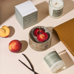 A styled flat-lay featuring a candle product with its box, a candle in a glass jar, a notebook, a book, sliced plums in a stone dish, vanilla beans, and a peach on a warm beige surface with soft shadows. The candles are labelled with the text 'Notebook' while the packaging box shows the company branding.