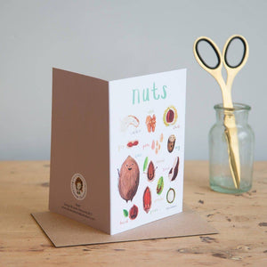Greeting card featuring illustrations of nuts on a white background. The heading 'nuts' is printed at the top of the card while each of the images are labelled with smaller text. The card stands on top of a brown envelope on a wooden surface beside a jar containing a scissors.