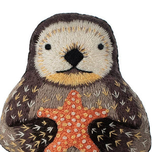 Close up showing the upper half of an embroidered otter plush doll with black, brown, grey, yellow, orange and white thread detailling, on a white background. 