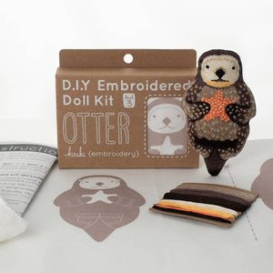 A DIY embroidered doll kit box next to a finished embroidered otter plush  doll on a white background. The box is brown with white text and sits behind an embroidery pattern and some threads. 