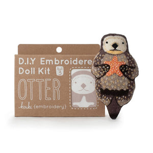 A DIY embroidered doll kit box next to a finished embroidered otter plush  doll holding a star, on a white background. The box is brown with white text. 