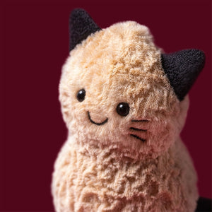 A close up image of a brown plush peanut shaped toy with black cat ears, eyes and whiskers on a wine coloured background. 