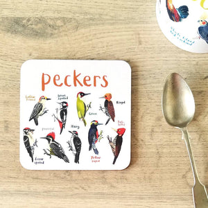 Coaster featuring illustrations of birds on a white background. Each bird is labelled, while the text 'Peckers' is printed on the top. The coaster sits on a wooden surface beside a spoon.
