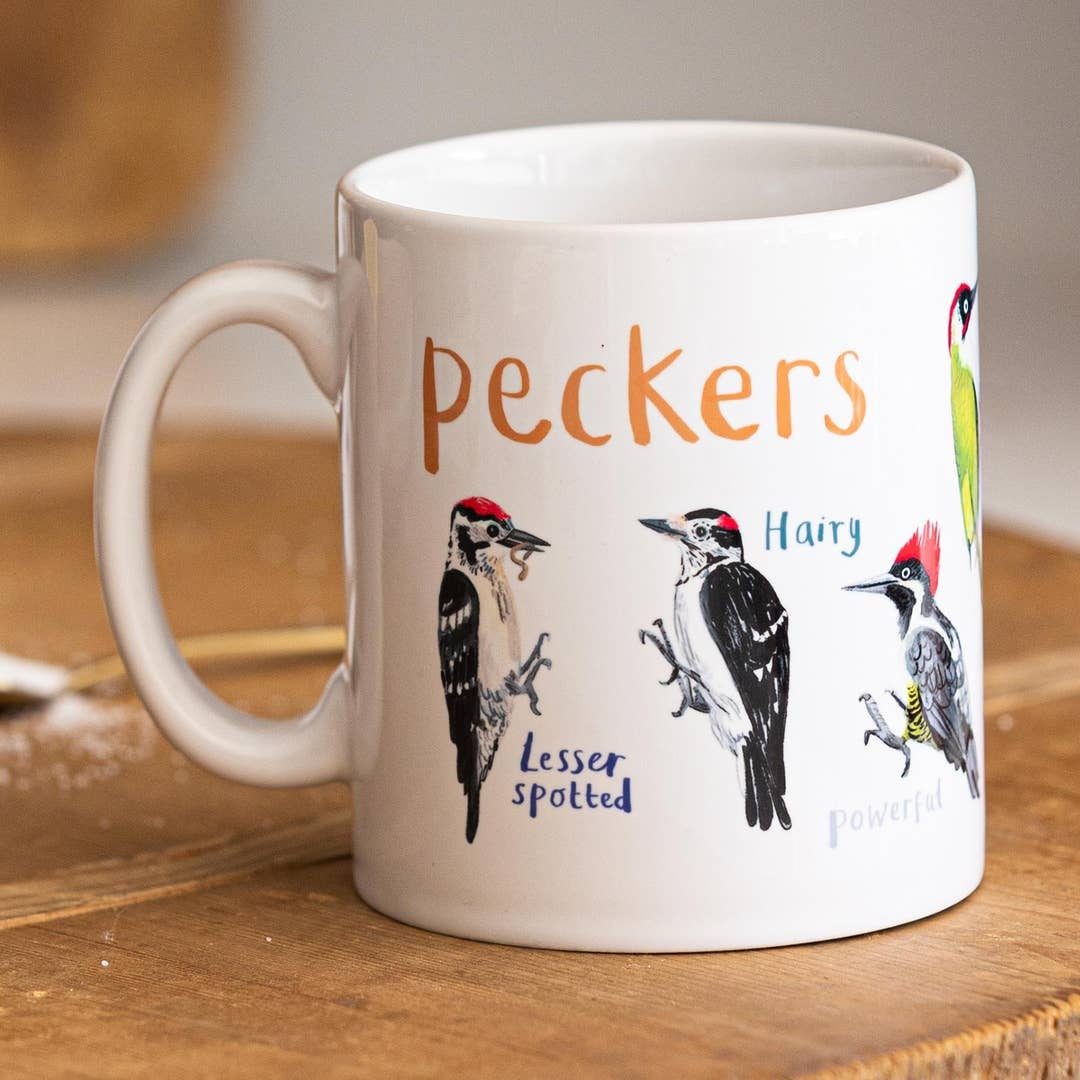 A white mug with colourful illustrations of different types of birds. The text 'Peckers' is printed over the images along with text labelling each image. The mug sits on a wooden surface. 