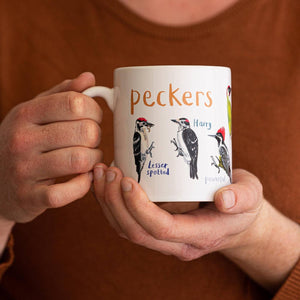 A person holding a white mug with colourful illustrations of different types of birds. The text 'Peckers' is printed over the images along with text labelling each image. 