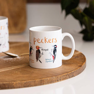 A white mug with colourful illustrations of different types of birds. The text 'Peckers' is printed over the images along with text labelling each image. The mug sits on a wooden surface. 