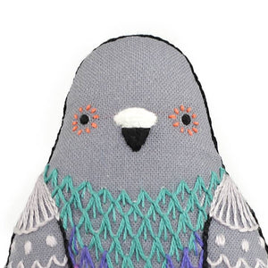 Close-up of a grey embroidered pigeon plush doll with black, white, orange and blue thread detailling. 