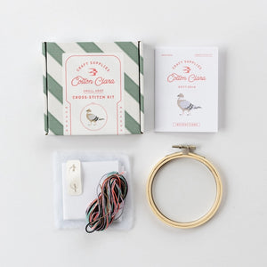 Cross-stitch kit packaging beside an instructions booklet, a wooden embroidery hoop and coloured threads on a white background.