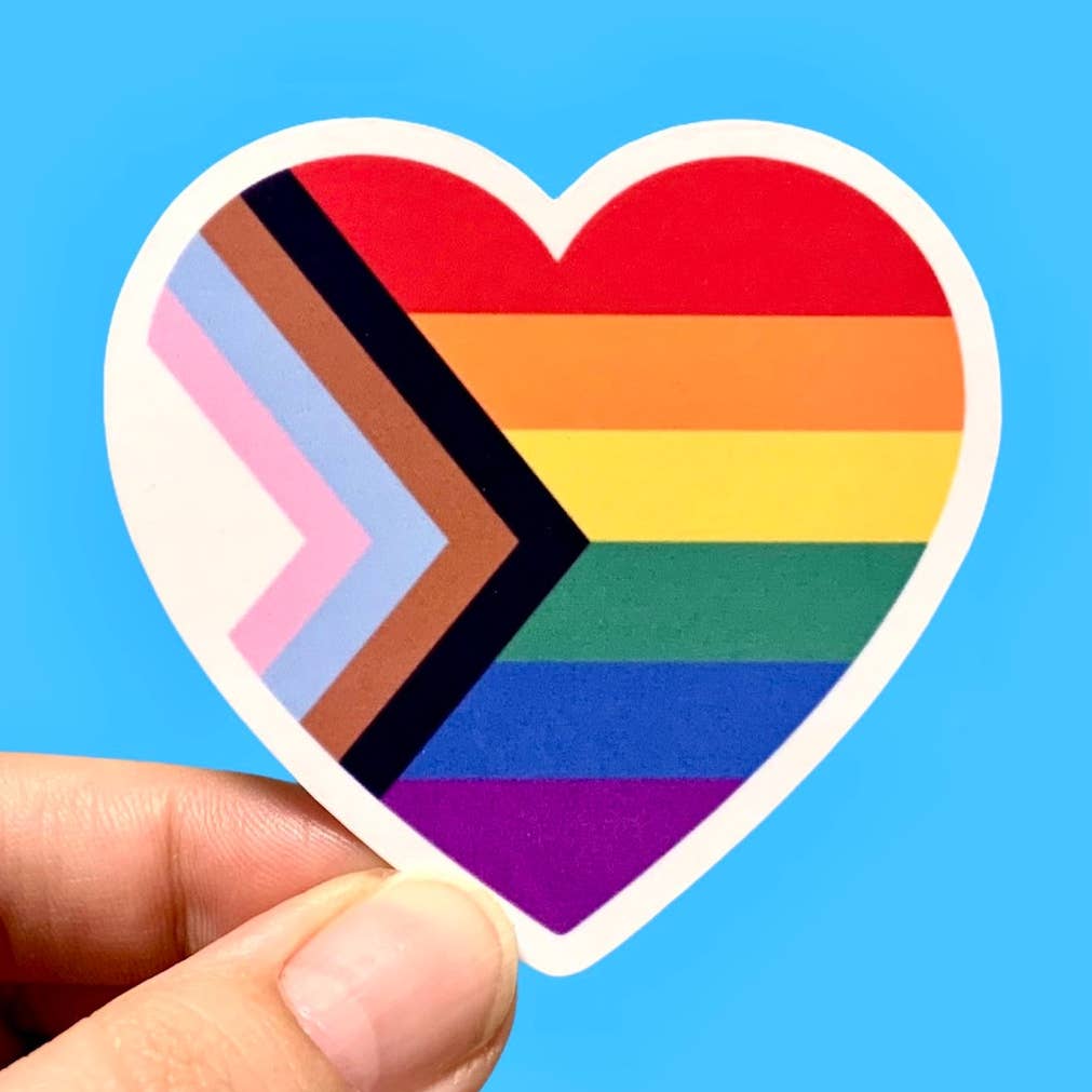 A heart-shaped sticker with a white border and a Pride flag design inside it. The sticker is held against a blue background. 