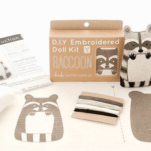 A DIY embroidered doll kit box next to a finished embroidered raccoon doll plush on a white background. The box is brown with white text and sits behind a raccoon embroidery pattern and some threads. 