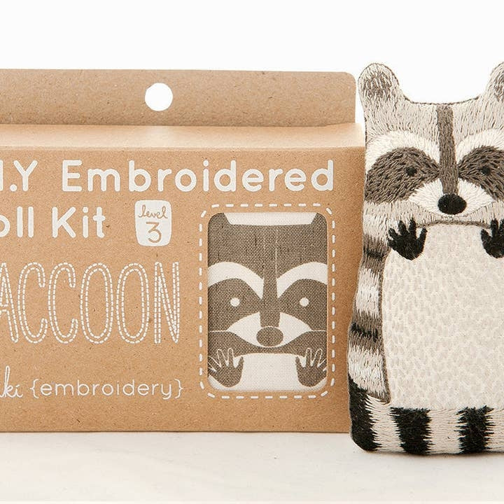 A DIY embroidered doll kit box next to a finished embroidered raccoon doll plush on a white background. The box is brown with white text. 