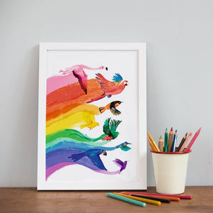 Art print featuring a colourful illustration of birds flying and forming a rainbow behind them on a white background. The print is in a white frame and stands on a wooden surface beside a container filled with coloured pencils. 