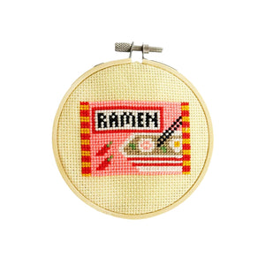 Cross-stitched design of a ramen pack in a wooden hoop on a white background.