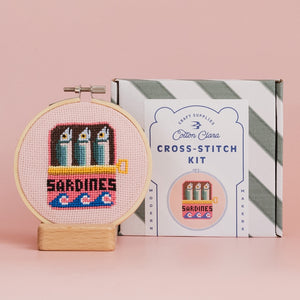 A cross-stitched design of sardines in a tin framed in a wooden hoop stands beside a packaging box with branding on a pink background. 