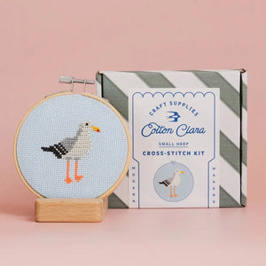 Cross-stitch kit with a completed seagull design contained in a circular embroidery hoop and Cotton Clara packaging on a pink background.