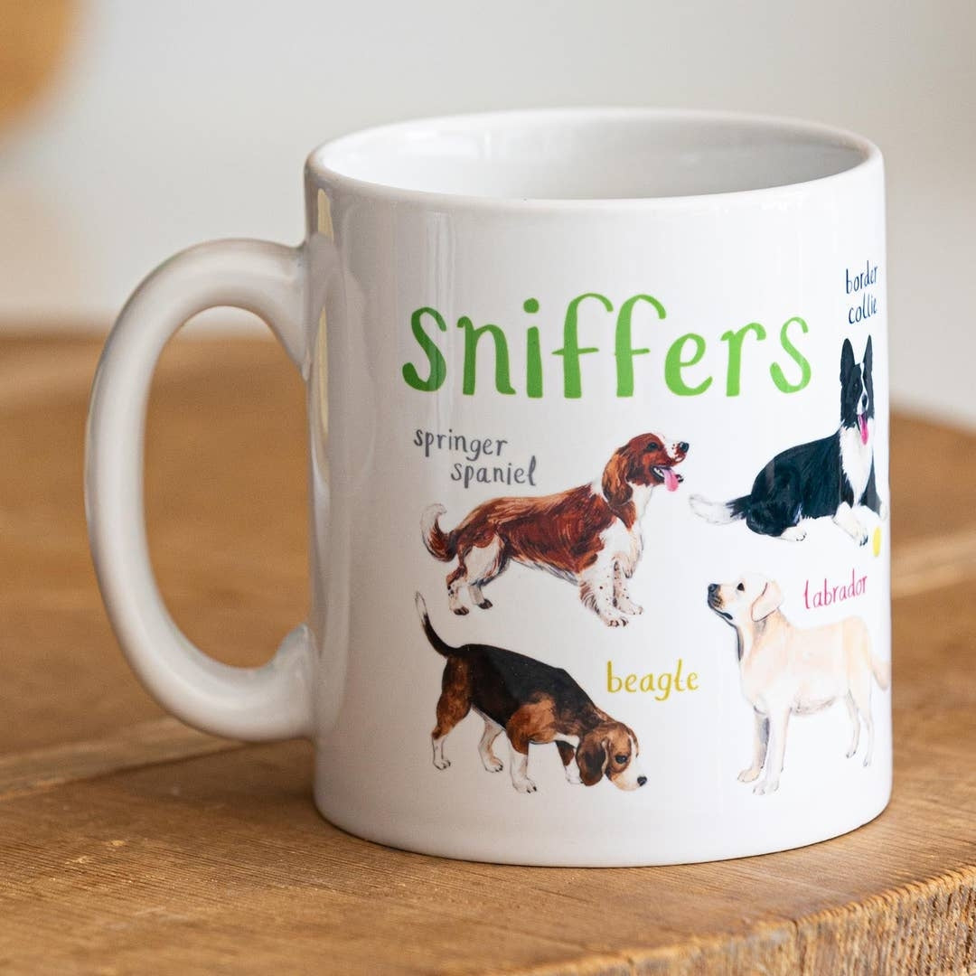 A white mug with dog illustrations. The text 'Sniffers' is printed over the illustrations along with text labelling each image. The mug sits on a wooden surface.