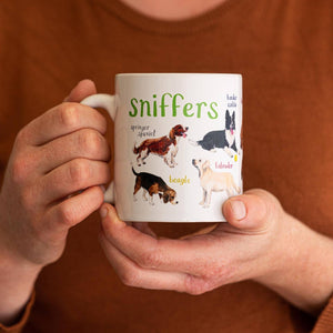 Person holding a white mug with dog illustrations. The text 'Sniffers' is printed over the illustrations along with text labelling each image. 