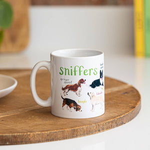 A white mug with dog illustrations. The text 'Sniffers' is printed over the illustrations along with text labelling each image. The mug sits on a wooden surface.