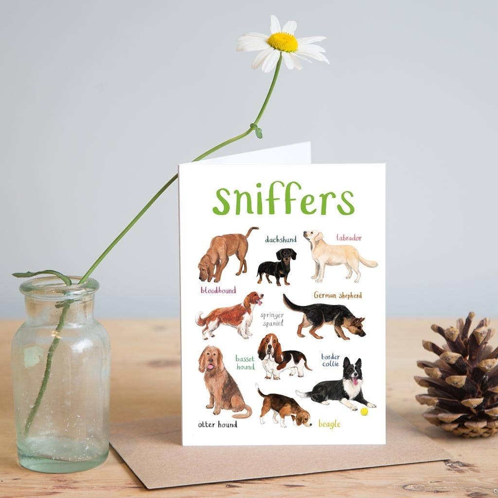 Greeting card featuring illustrations of dogs on a white background. The heading 'sniffers' is printed at the top of the card while each of the images are labelled with smaller text. The card stands on top of a brown envelope on a wooden surface. A jar containing one flower and a pine cone sit on either side of the card. 