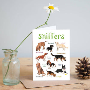 Greeting card featuring illustrations of dogs on a white background. The heading 'sniffers' is printed at the top of the card while each of the images are labelled with smaller text. The card stands on top of a brown envelope on a wooden surface. A jar containing one flower and a pine cone sit on either side of the card. 