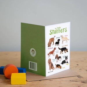 Greeting card featuring illustrations of dogs on a white background. The heading 'sniffers' is printed at the top of the card while each of the images are labelled with smaller text. The card stands on top of a brown envelope on a wooden surface. Three colourful blocks sit beside the card. 