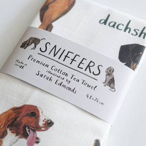 A folded white cotton tea towel showing dog illustrations with text printed underneath. A paper label with the text 'Sniffers' is wrapped around the middle of the towel, along with the text 'Premium Cotton Tea Towel', 'illustrated by Sarah Edmonds' and 'made in the UK'.