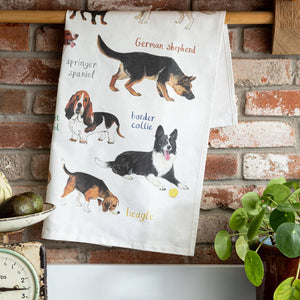 A white cotton tea towel showing dog illustrations with text printed underneath. The towel is folded and hangs from a wooden dowel against a red brick wall background. 