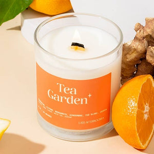 A candle in a milky-white glass jar filled with white wax and a lit wick. The text 'Tea Garden' is printed on an orange label on the candle along with the company branding. The candle sits on a surface beside half of an orange and some ginger. 