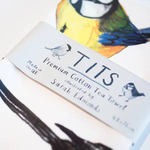 Tea towel with a colourful bird illustration. A paper label is wrapped around the middle of the tea towel, with the text 'Tits' and 'Premium Cotton Tea Towel' printed on it. 