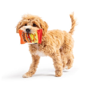 Image of a dog carrying a toy in it's mouth. The toy is designed to look like a bag of Cheetos. 
