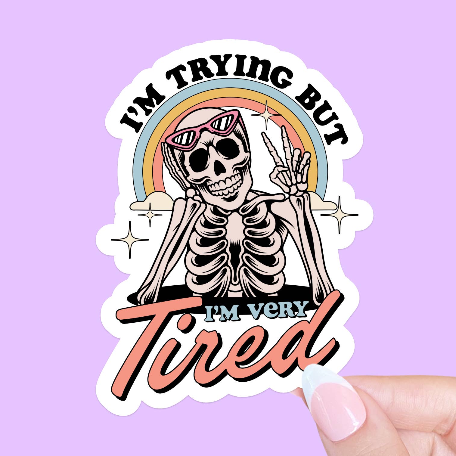A sticker featuring an illustration of a skeleton wearing sunglasses and using their fingers to give a peace sign. A rainbow sits in the background, behind the skeleton along with two clouds. The text 'I'm trying but I'm very tired' is also printed around the skeleton. The sticker is held against a purple background. 
