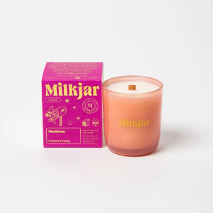 Canadian company Milkjar candle and its box packaging. The white candle is in an orange glass with "Milkjar" written in yellow. The other side of the candle has a cute mailbox doodle in yellow. The packaging is a small pink box with yellow text and graphics. 
