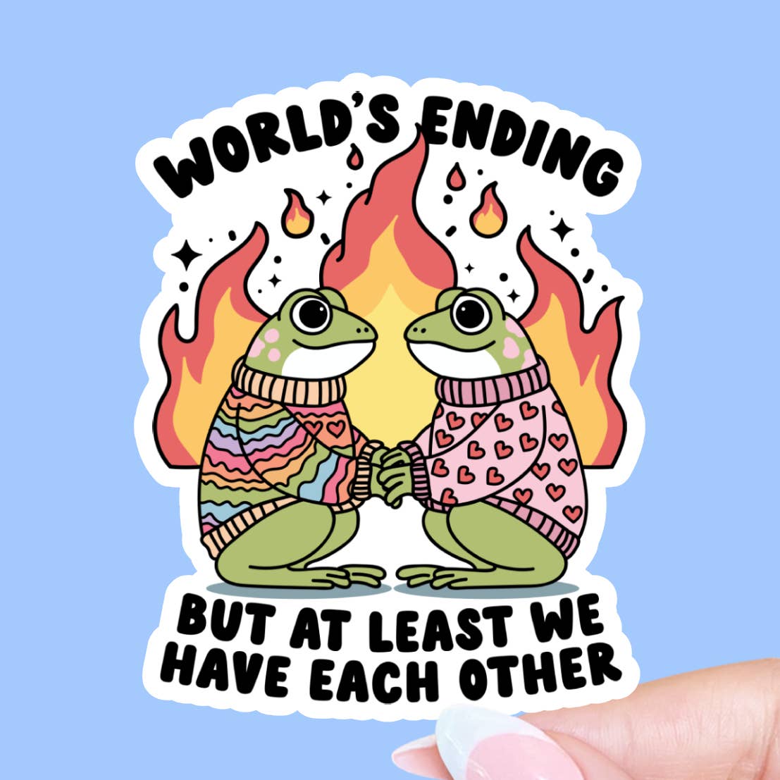 A sticker featuring illustrations of two frogs wearing sweaters and holding hands as they gaze into each other's eyes. The text 'World's Ending But At Least We Have Each Other' is printed around the frogs while flames burn behind them. The sticker is being held against a blue background. 
