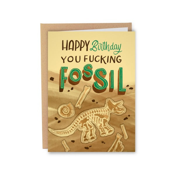 yellow birthday card with dirt dunes and bones (shell, triceratop, stray bones). top text reads "happy birthday you fucking fossil"