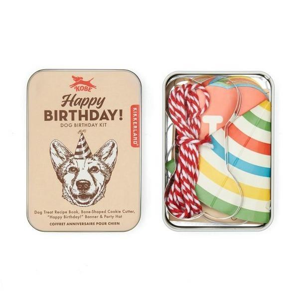 open metal container. the lid is beige with an illustration of a smiling dog with a party hat. top text reads "happy birthday! dog birthday kit". opened container reveals a bone cookie cutter, a party hat, and more. kikkerland product on white background.